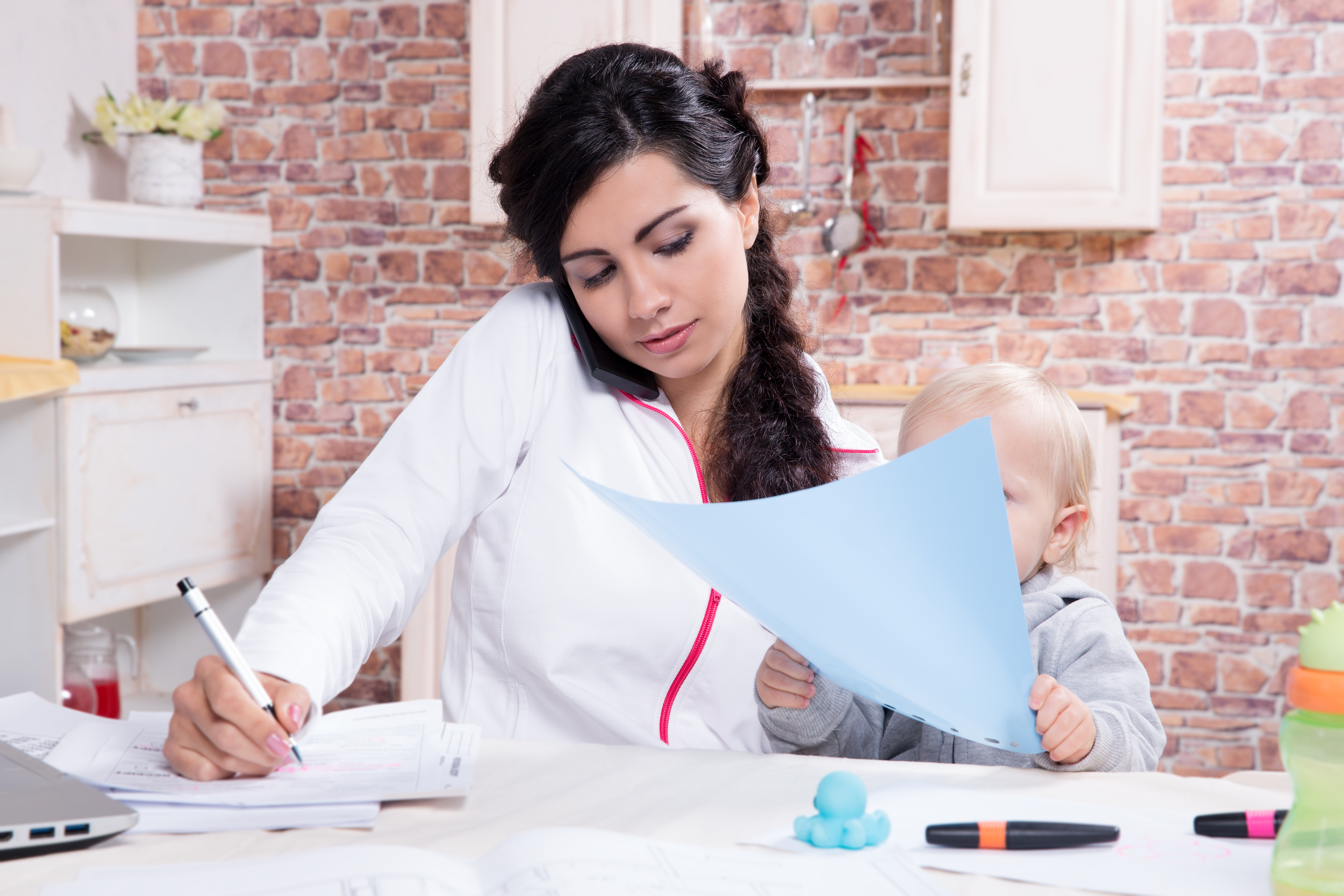 Mother with baby in the kitchen working with documents and speaks by phone